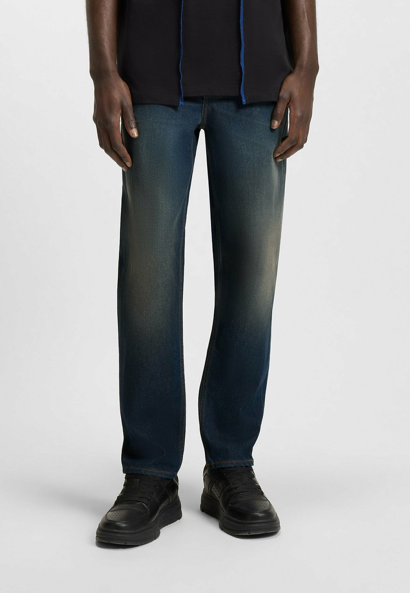 Jeans in denim blu con taglio dritto, caratterizzati da una finitura scolorita, cuciture evidenziate e texture usurata, abbinati a sneaker nere.