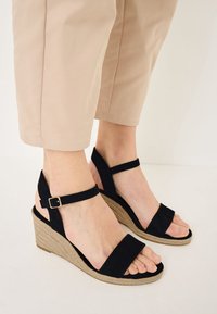 Sandales compensées en daim noir avec larges brides et boucle à la cheville, dotées d'une base tissée texturée. Le design comprend des orteils et des talons ouverts.