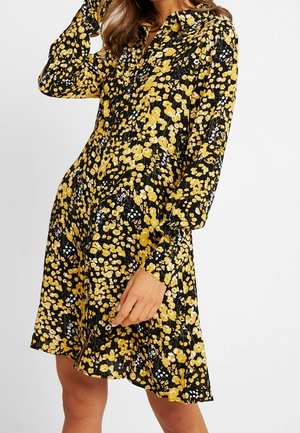 Femme portant une robe noire à manches longues avec un motif floral jaune, debout devant un fond uni.