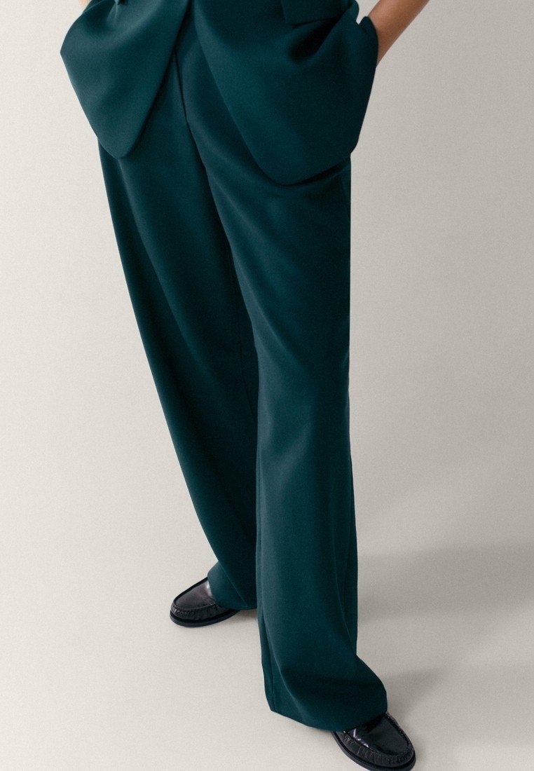 Personne portant un pantalon ample vert foncé avec de grandes poches avant et des mocassins noirs, debout les mains dans les poches.