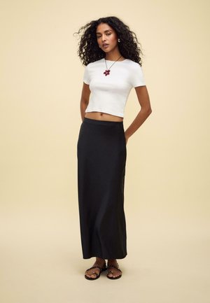 Jupe noire longue jusqu'au sol associée à un haut ajusté à manches courtes blanc. Accessoirisée avec un collier de pendentif rouge et des sandales noires.