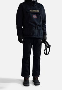 Personne portant une veste d'hiver noire avec un patch du drapeau norvégien, des gants noirs, un pantalon de neige, des bottes, et tenant des bâtons de ski noirs.