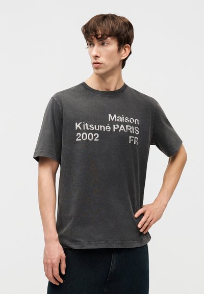 Νεαρός άνδρας με κοντά καστανά μαλλιά, φορώντας μια ξεθωριασμένη μαύρη μπλούζα Maison Kitsuné Paris 2002 FR με τύπωμα, στέκεται με το ένα χέρι στη μέση.