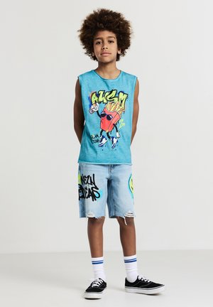 Junge mit lockigem Haar, der ein blaues ärmelloses Shirt mit einem Cartoon-Pommes-Design und eine Jeansshorts mit Graffiti-Drucken trägt.