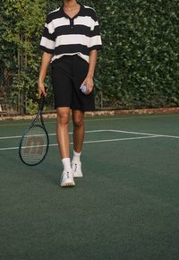 Polo a righe bianco e nero, pantaloni corti neri, sneakers bianche con dettagli, che tiene una racchetta da tennis e una palla blu, su un campo da tennis.