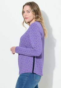 Fioletowa bluzka z długim rękawem z kwiatowym wzorem, wykonana z miękkiego materiału o luźnym kroju oraz z rozcięciem na dole. W komplecie z niebieskimi dżinsami.