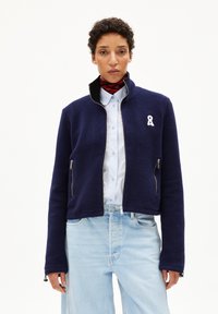 Veste en polaire bleu marine avec fermeture éclair à l'avant, deux poches et un logo en ruban blanc. Elle présente un col structuré et une coupe ajustée.