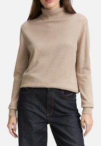 Pull en col roulé beige en tissu tricoté doux, avec des manches longues et un col côtelé, associé à un jean en denim foncé avec des coutures visibles.