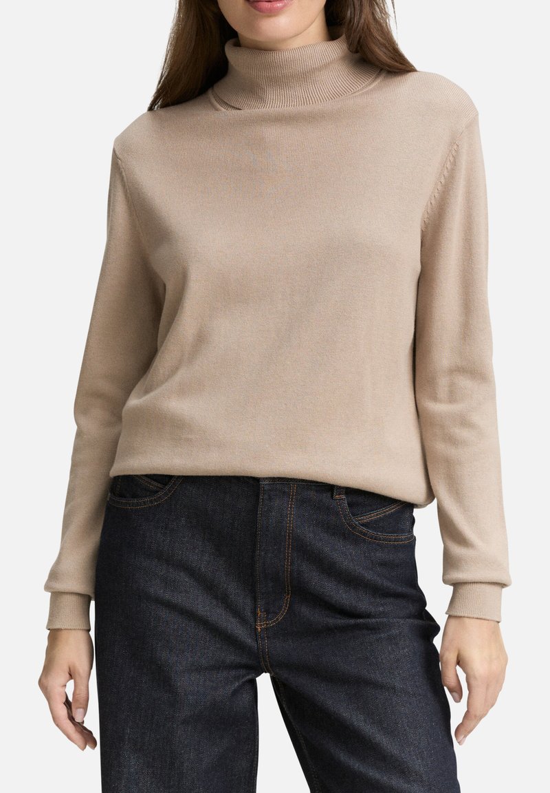 Pull en col roulé beige en tissu tricoté doux, avec des manches longues et un col côtelé, associé à un jean en denim foncé avec des coutures visibles.