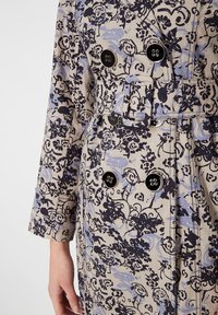 Abrigo de tela clara con intrincados patrones florales en azul marino y azul claro, que cuenta con botones de doble botonadura y un cinturón en la cintura.