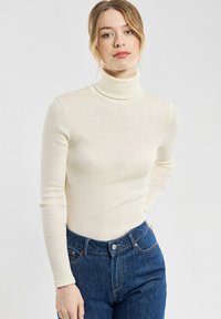 Cremfarbener Rollkragenpullover aus geribbeltem Stoff, mit langen Ärmeln und taillierter Passform, kombiniert mit dunkelblauen hochgeschnittenen Jeans.