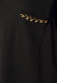 Top de tela negra con un bolsillo que presenta un detalle de cadena dorada. Textura suave con un diseño sencillo y costuras visibles en los bordes.