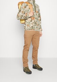 Giacca in pile con motivo floreale verde e arancione, pantaloni cargo beige e scarpe da escursionismo nere con lacci gialli; zaino giallo.