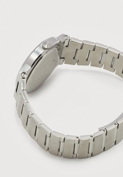 Ασημένια μεταλλική μπάντα ρολογιού και πίσω μέρος κάσας με βραχιόλι συνδέσμων, τοποθετημένα επίπεδα σε λευκή επιφάνεια, που δείχνουν πλευρική όψη και λεπτομέρειες κλειδώματος.