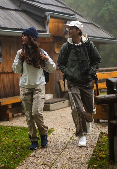 Two individuals walk on a stone path. One wears a gray sweatshirt, beige cargo pants, and navy shoes; the other sports a green jacket, beige pants, and white shoes.