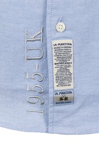 Ljusblå tygskjorta med vit knapp, broderad med "1955-UK" och en etikett som visar varumärkesinformation och vårdinstruktioner.