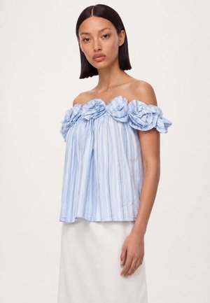Femme aux cheveux raides et foncés porte un haut rayé bleu et blanc à épaules dénudées avec de grandes fleurs en tissu le long de l'encolure et une jupe blanche.