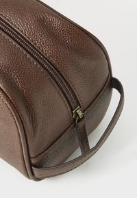 Bolsa de toalete em couro castanho texturizado, com fecho de zíper e pega, em fundo branco.