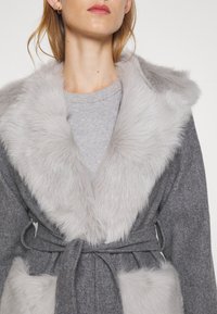 Woman wearing a gray coat with large light gray faux fur collar and pocket trims, tied at the waist over a light gray knit top.