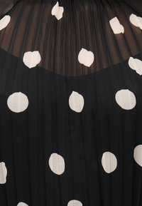 Μαύρο πλισέ ύφασμα με μεγάλα λευκά πουά, με διαφανή κορυφαία ενότητα που έχει το ίδιο μοτίβο πουά.