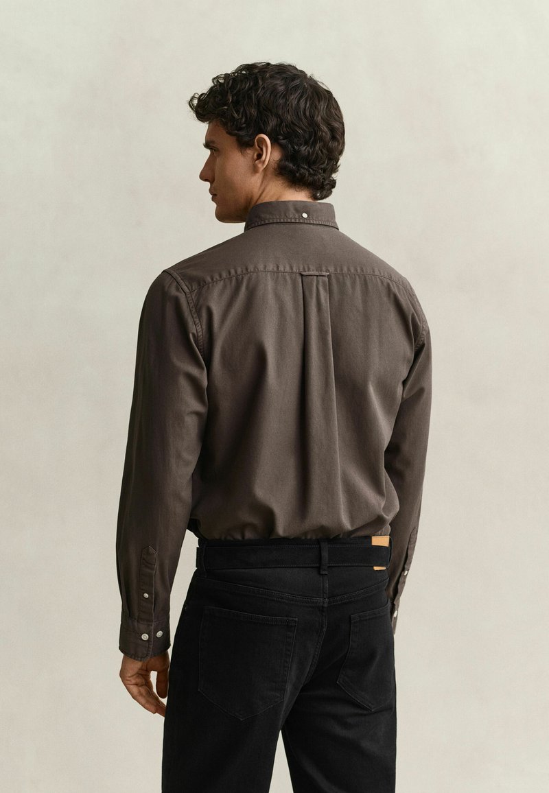 Mann mit lockigem dunklem Haar trägt ein dunkelbraunes, langärmliges Hemd mit Knöpfen, das in schwarze Jeans gesteckt ist, und steht mit dem Rücken zu einem schlichten Hintergrund.