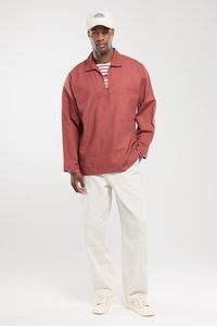 Rode geweven pullover met een V-halsontwerp en voorzak. Gecombineerd met lichtgekleurde wijduitlopende broeken en crèmekleurige sneakers. Licht petje op.