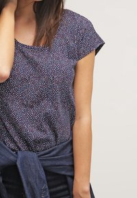 Mujer que lleva una blusa corta de mangas con patrón en azul marino y una chaqueta de mezclilla atada a la cintura, de pie contra un fondo neutro.