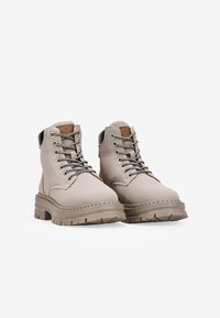 Bottes à chevilles beiges en cuir lisse avec une semelle en caoutchouc épaisse et texturée, un design à lacets et des accents de talon noirs contrastants.
