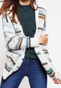 Stickad cardigan i vitt med flerfärgade ränder, med lös passform, öppen front och ribbad textur. Bärs över en mörkgrön topp.