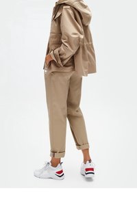 Persona che indossa un giubbotto e pantaloni beige con scarpe bianche con accenti rossi e neri, in piedi con le mani in tasca.