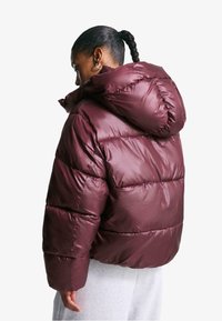 Bordeaux pufferjack met een capuchon, met een glanzende afwerking, geborduurd ontwerp en elastische manchetten. De achterkant toont een ontspannen pasvorm.