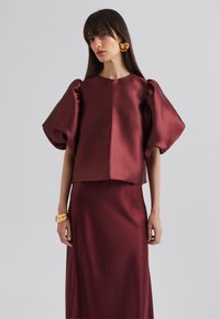 Haut en satin bourgogne avec des manches bouffantes volumineuses, associé à une jupe longue assortie. Présente une texture lisse et un design minimaliste.