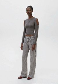 Maglietta grigia a maniche lunghe con design a spalle scoperte, abbinata a jeans larghi grigi chiari e scarpe rosse, su sfondo neutro.