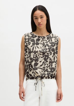 Jeune femme aux longs cheveux foncés portant un haut sans manches noir et blanc à motif floral avec lien à la taille et un pantalon blanc, fond neutre.