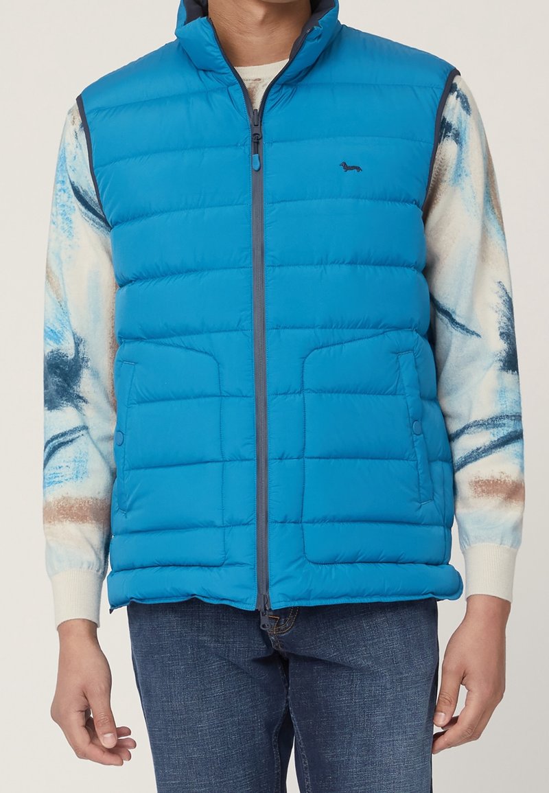 Mand iført en klar blå quiltet ærmeløs dunvest over en hvid og blå mønstret langærmet skjorte med mørkeblå jeans.