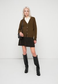 Jovem mulher de pé, vestindo um cardigan castanho com botões, saia escura, botas pretas até ao joelho, camisa branca e a carregar uma bolsa preta de ombro.