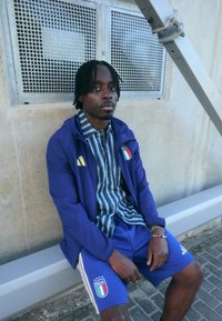 Adidas trenirka sadrži plavu jaknu s talijanskom zastavom i žutim detaljima, u kombinaciji s plavim šorcem. Ispod se nosi prugasta majica. Betonska pozadina.