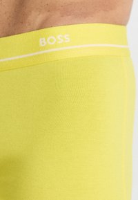 Gule boxer briefs lavet af strækbart materiale, med en hvid talje med "BOSS" branding og glat, tekstureret stof.