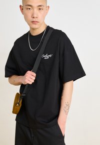 Crna pamučna t-shirt oversized kroja s bijelim logotipom "Carhartt WIP". Model nosi žutu torbicu preko tijela i nosi srebrnu lančić ovratniku.