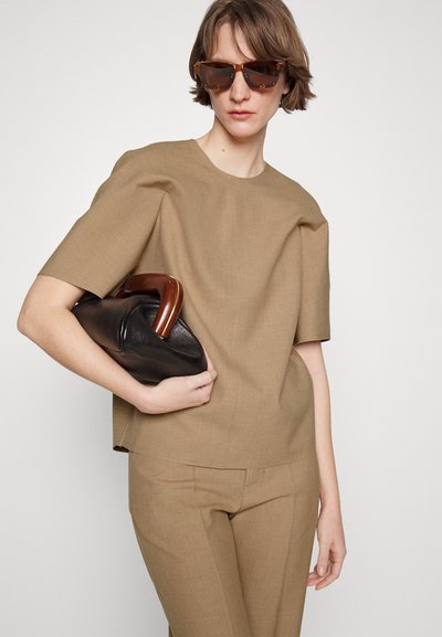 Bež lühikeste varrukatega topp, sirge lõike ja tekstuurse kangaga, koos sobivate kitsaste pükstega. Hoides musta nahast kotti puidust käepidemega.
