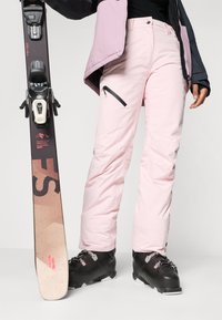 Skiausrüstung mit pinkfarbenen wasserdichten Hosen mit Reißverschlusstasche, schwarzen Skischuhen und einem dunklen Snowboard mit Logo und Farbverlauf.