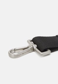 Cintura in tessuto nero con una chiusura in metallo argentato, con il marchio prominente "TOMMY HILFIGER." Superficie testurizzata, forma rettangolare e design robusto.