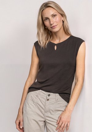 Kvinde med blondt hår iført sort ærmeløs top og beige bukser, der står mod en lys baggrund med et let smil.