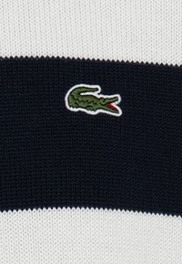 Stickad bomullspolo med horisontella marinblå och vita ränder. Broderad grön krokodil-logga med röda accenter på vänster bröst.