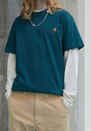 Personne aux cheveux blonds portant un t-shirt Carhartt bleu sarcelle sur un t-shirt blanc à manches longues, un pantalon en velours côtelé beige et un collier chaîne argenté.