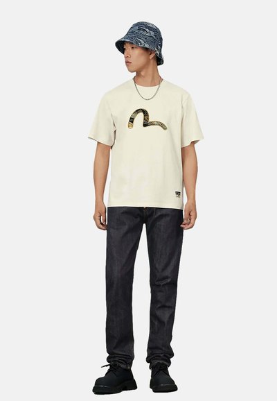 Maglietta crema a maniche corte con grafica di serpente nero e oro, abbinata a jeans denim scuro e cappello bucket blu con motivo. Scarpe nere.