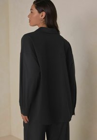 Black long-sleeve shirt made from a smooth fabric, featuring a classic collar, buttoned cuffs, and a curved hem for a relaxed fit.
