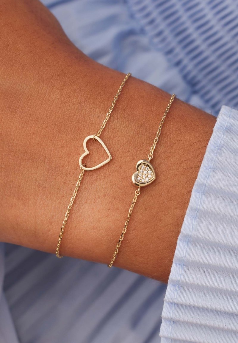 Goldene Armbänder in Herzform; ein offenes Herzdesign, das andere mit kleinen Steinen. Einfache Ketten, glatte Textur, am Handgelenk getragen.