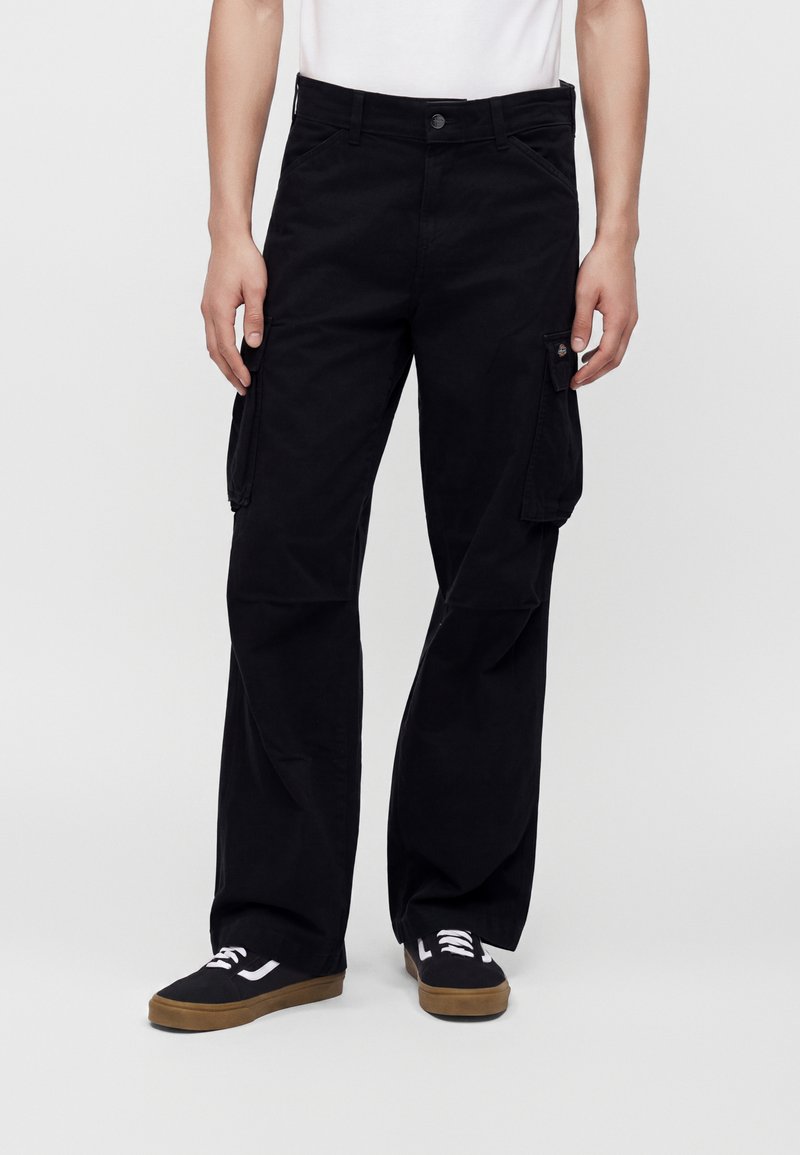 Personne portant un pantalon cargo noir et des baskets noires avec des lacets blancs et des semelles en gomme, debout devant un fond uni.