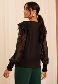 Top nero con maniche lunghe trasparenti caratterizzato da motivi a pois e floreali. Accenti arricciati sulle spalle e polsini testurizzati. Abbinato a pantaloni verdi.